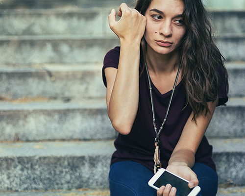 stock photo of a young woman sitting on outside steps holding mobile phone in one hand and crying