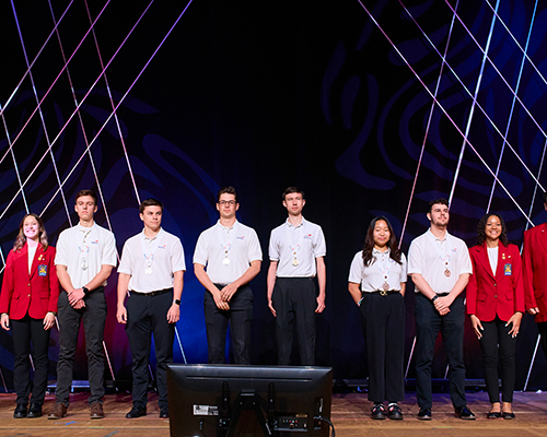 A small group of young men and women standing in a line wearing white shirts and black pants, some wearing red jackets