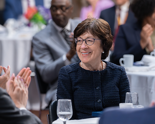Close-up of a smiling white woman seated at a dinner table with people seated at other tables in the background.