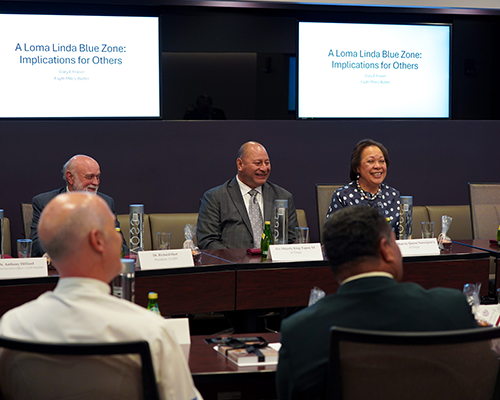 A photo of a smiling king and queen of Tonga sitting at a boardroom table in front of a slide on the Loma Linda Blue Zone. 