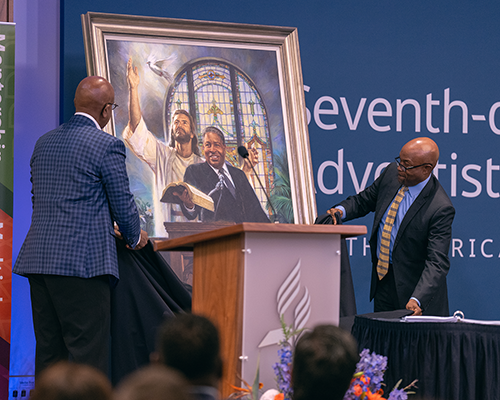 Two black men on a stage unveil a painting of a black man preaching 