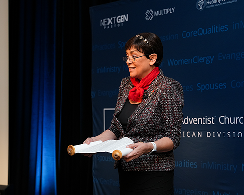 A Hispanic woman holding a scroll speaks at a conference.