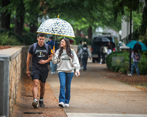 Young man and woman walking together under a flowered umbrella on college campus