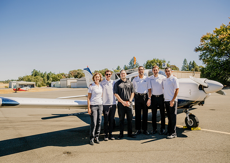 A diverse group of five men and one woman pose in front of an airplane. 