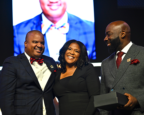 Black man, black woman, black man smiling on a stage. 
