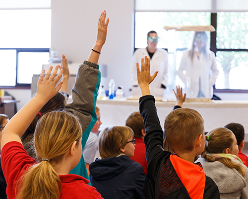 Back shot of students in a classroom with hands raised. 