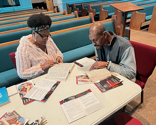 An African American woman studies the Bible at a table by a church pew with an African American man 