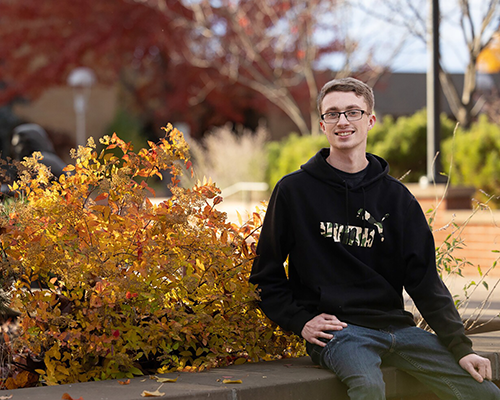 Bespectacled young man wearing a black hoodie sits on a stone wall in front of an orange bush. 
