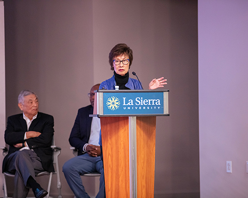 An Asian woman stands behind a podium, speaking, as an Asian man looks on.