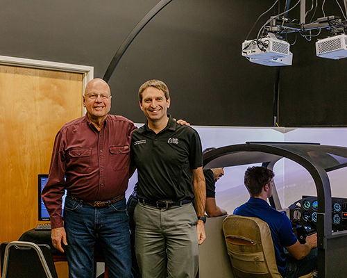 An older man and younger man stand together in an aircraft simulator. 