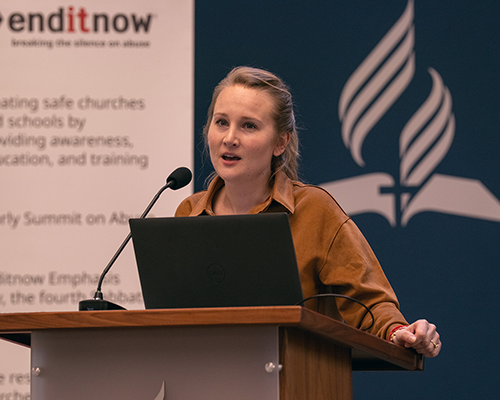 A white woman stands at a podium with enditnow poster behind her