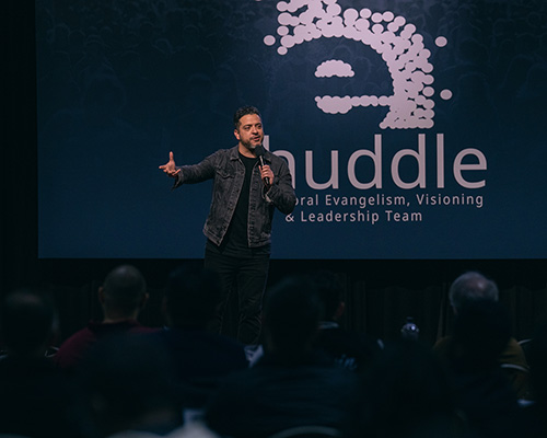Hispanic man speaks on a stage with eHuddle on a screen behind him.