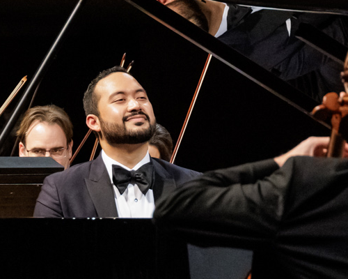 Jonathan Mamora plays a grand piano, accompanying an orchestra in front of a crowd of people. 