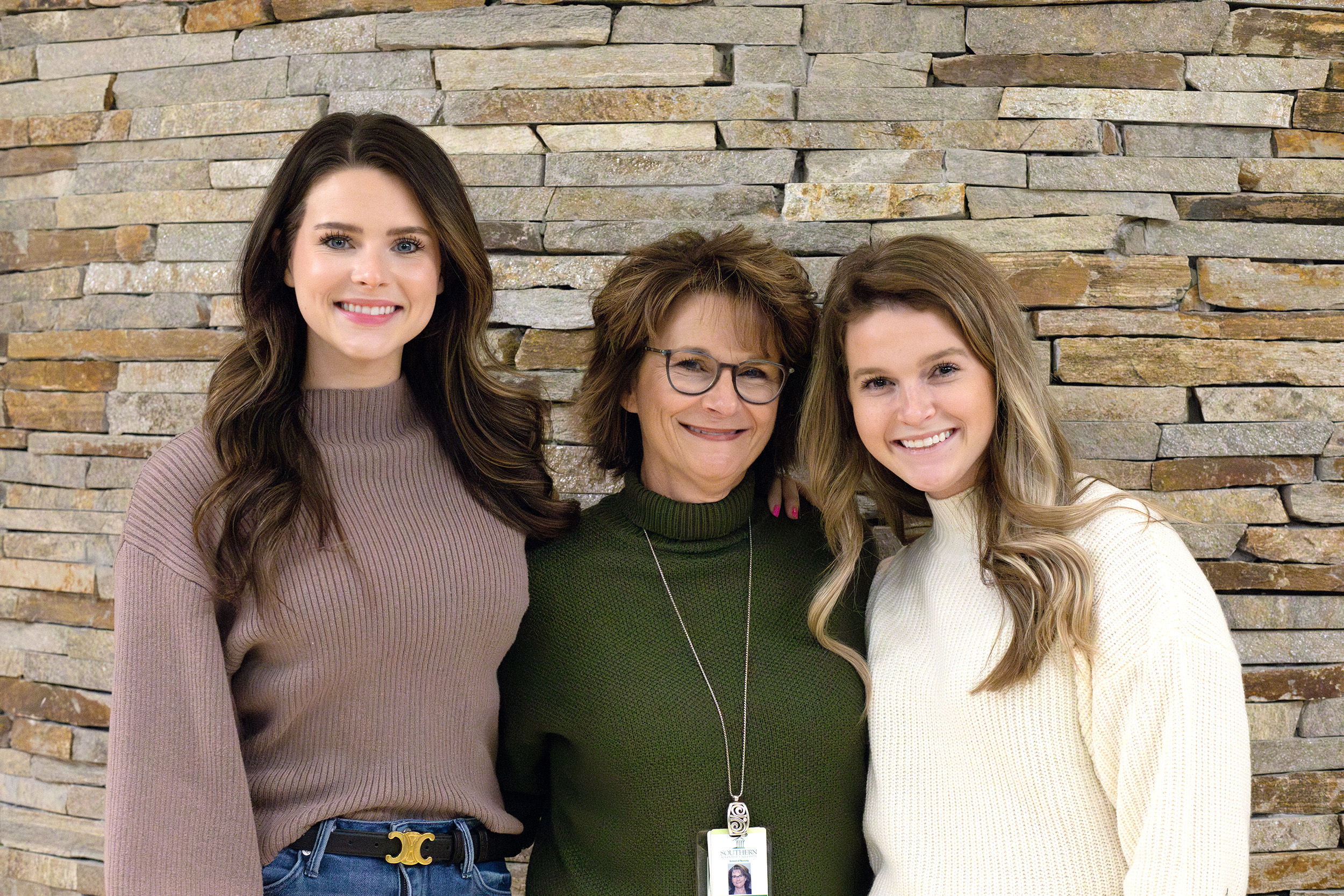 Three women, two brunette and one blonde, smiling towards the camera wearing sweaters.