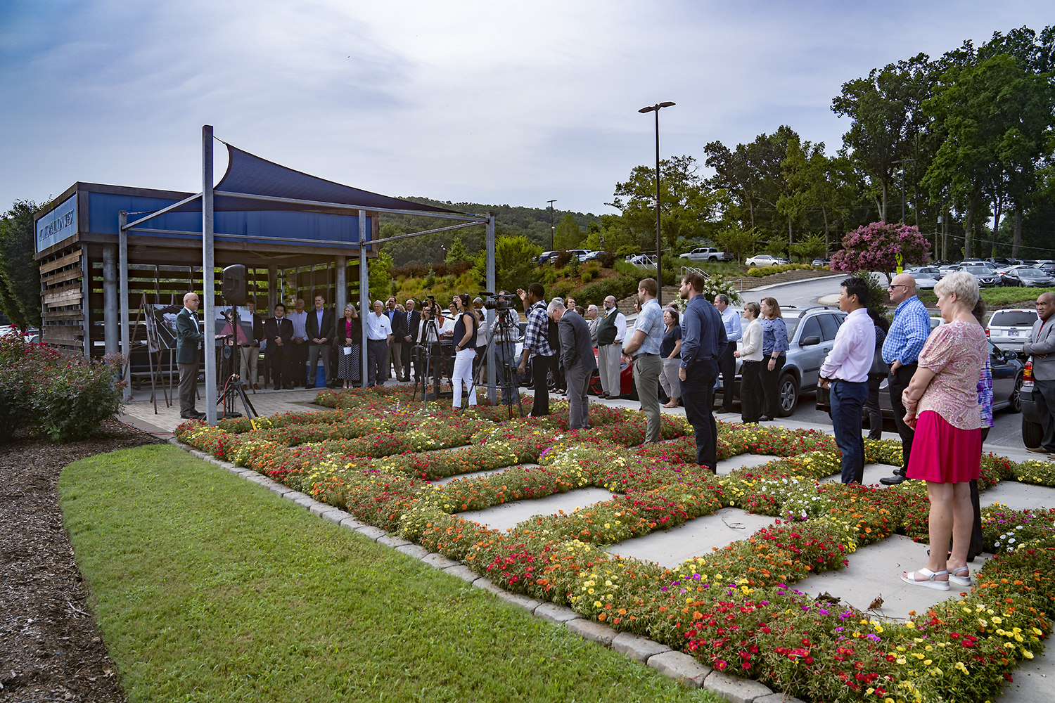 A man stands outside speaking under a tent while cameras record him and people stand listening.