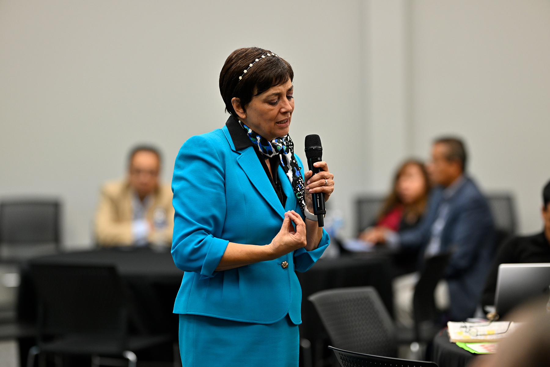 A Hispanic woman speaks in the middle of a room, holding a microphone. 