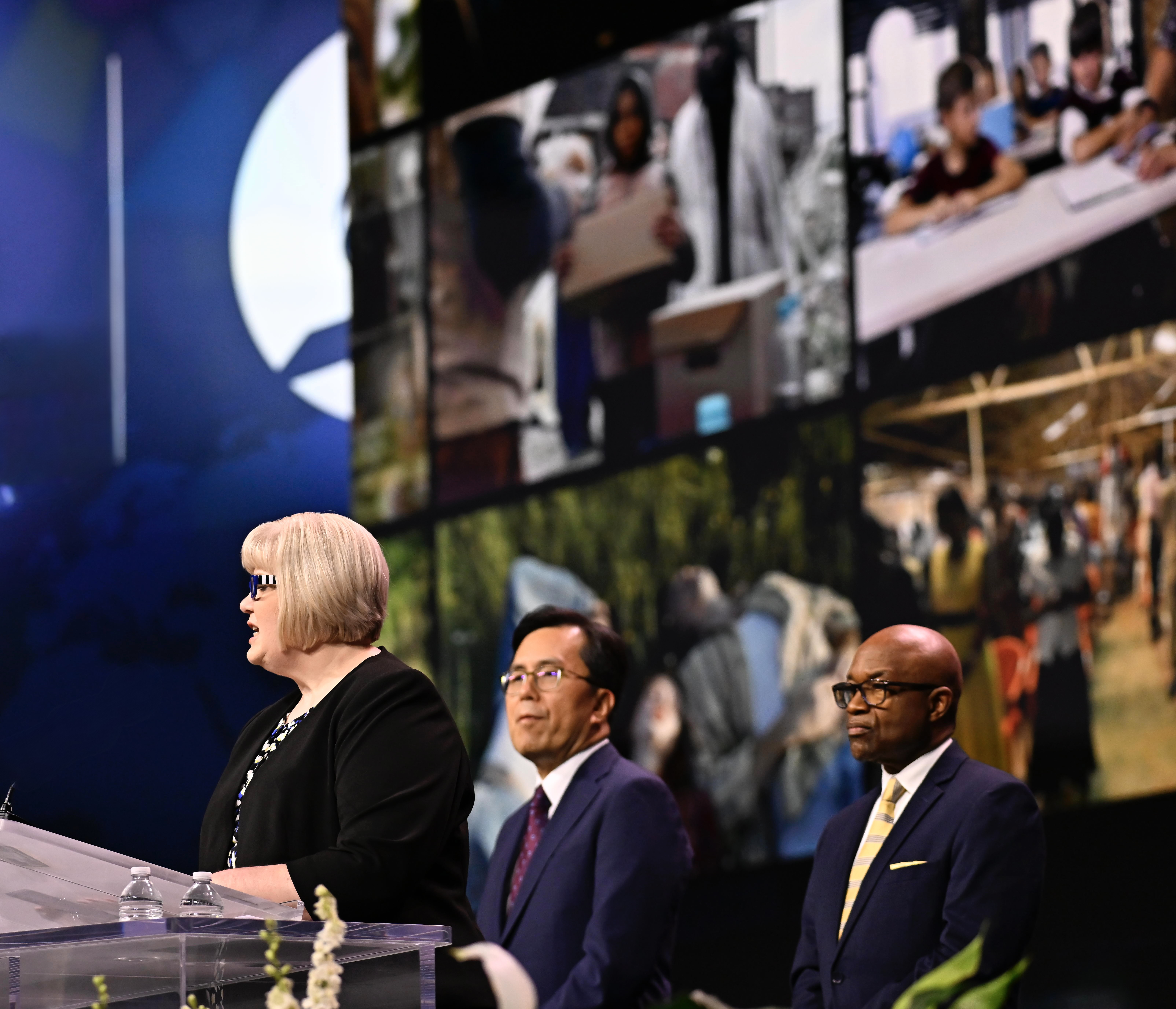 A blonde woman stands at a podium, speaking, as an Asian and black man watch her in the background.