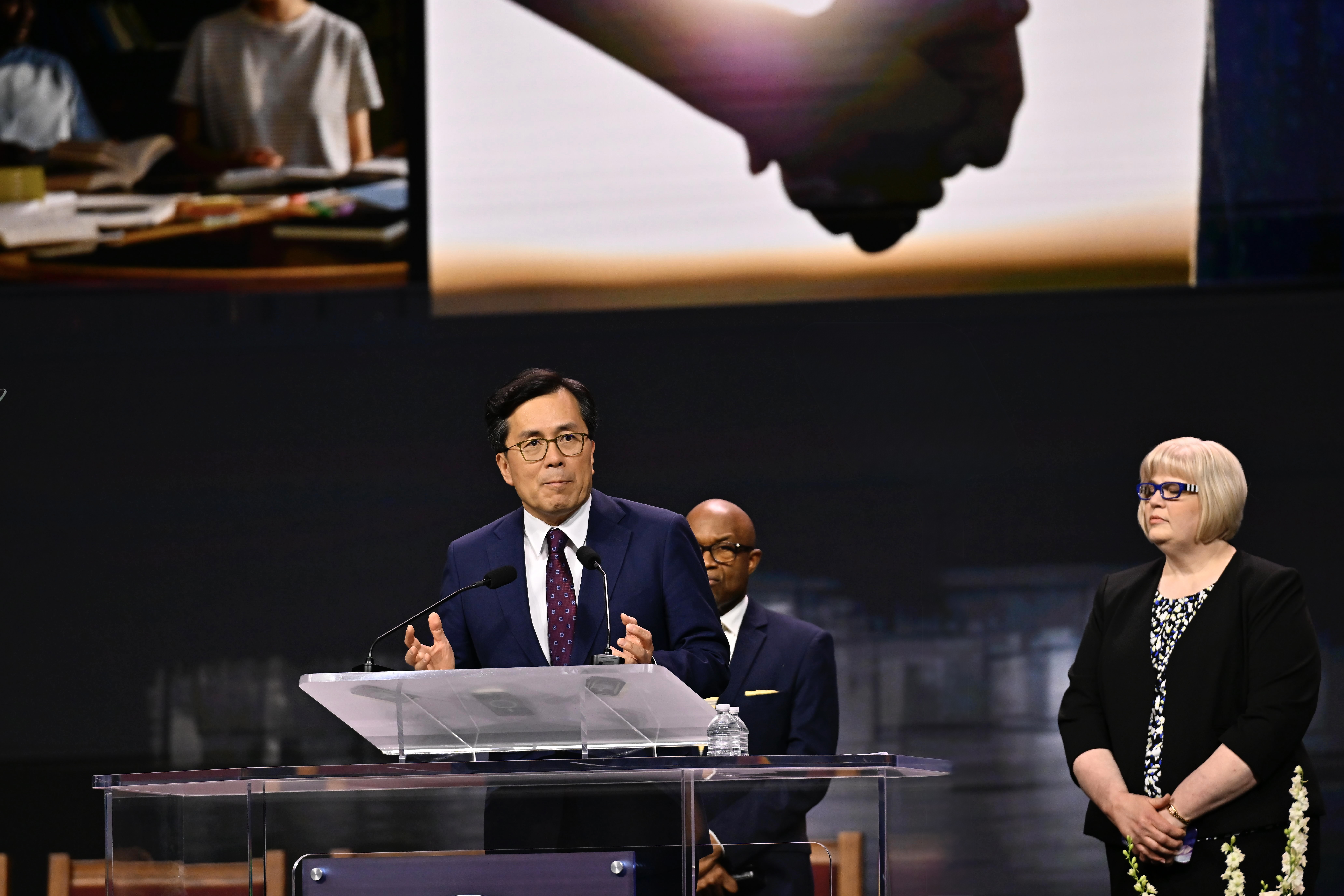 An Asian man speaks at a podium, with a black man and white woman in the background. 