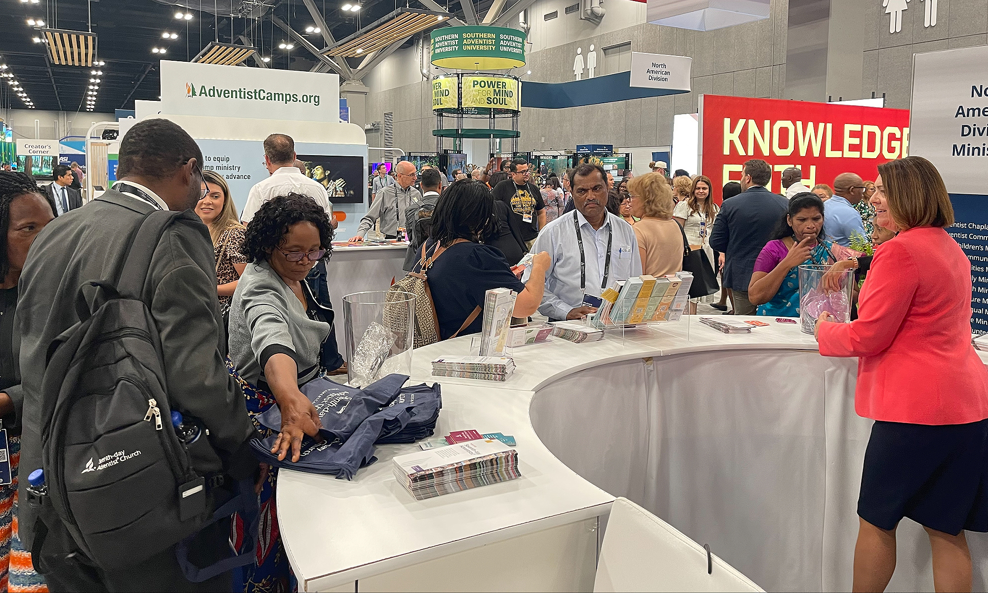 People crowd around a booth in a busy exhibit hall. 