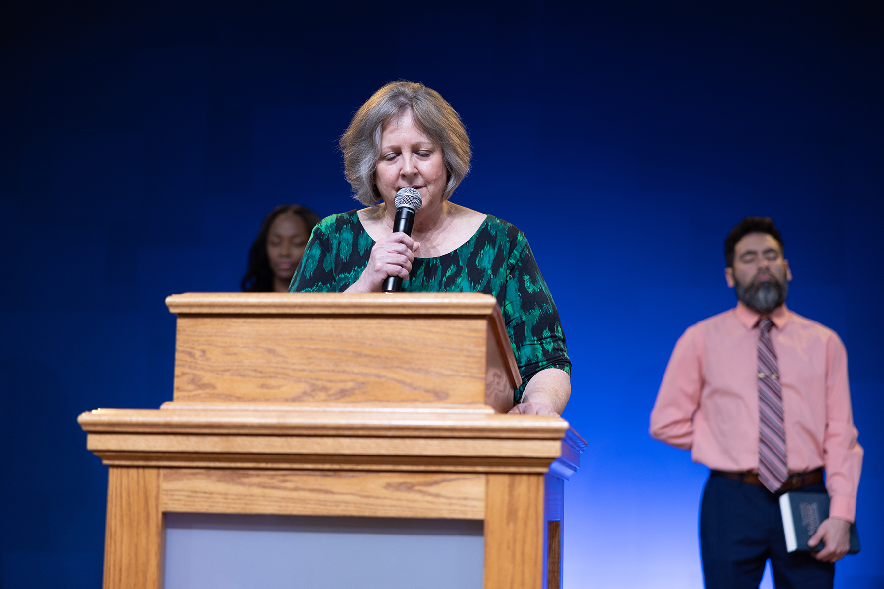 A woman standing behind a podium prays as a young woman and man stand on either side of her