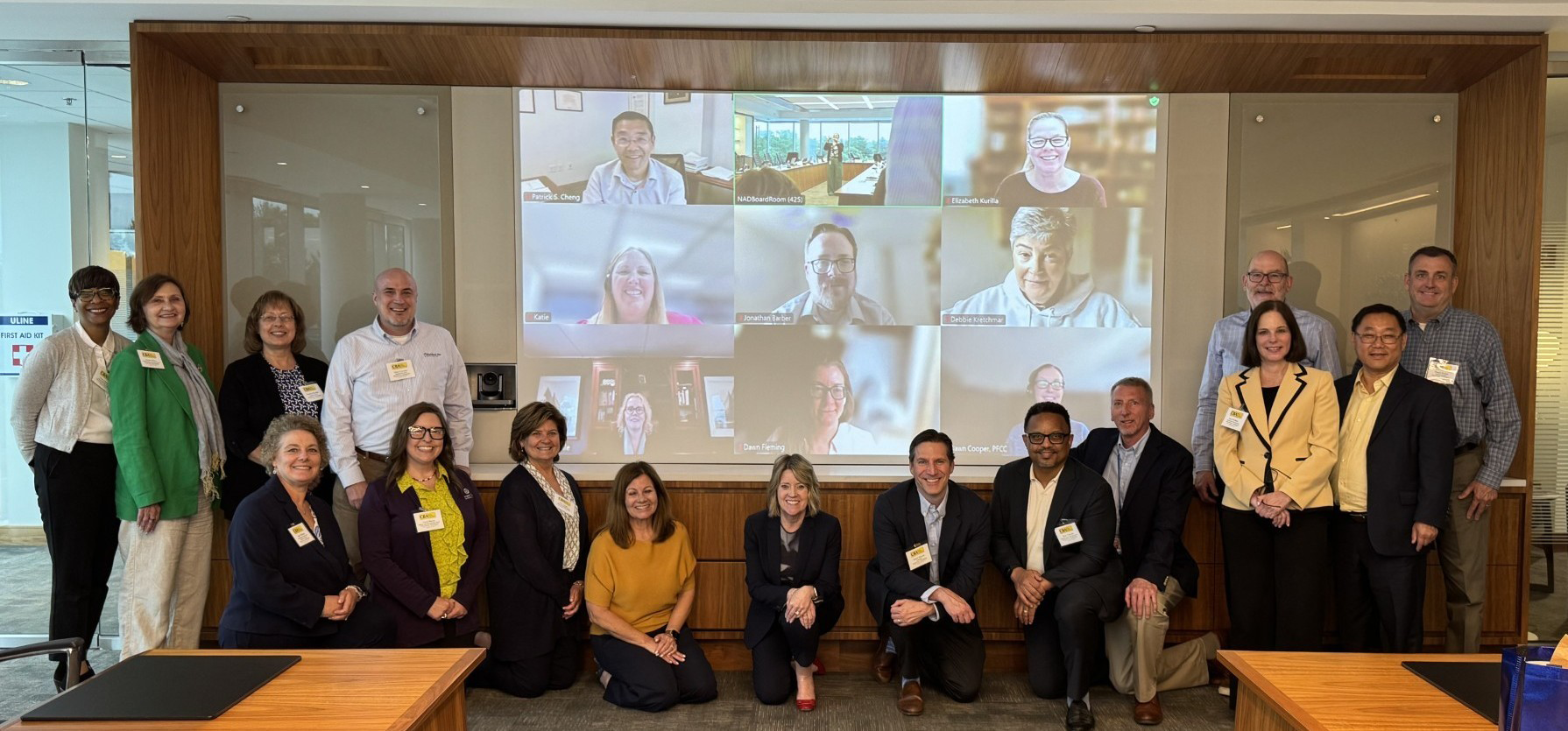 Photo of several people in a board room and many more on a screen in a Zoom meeting behind them.