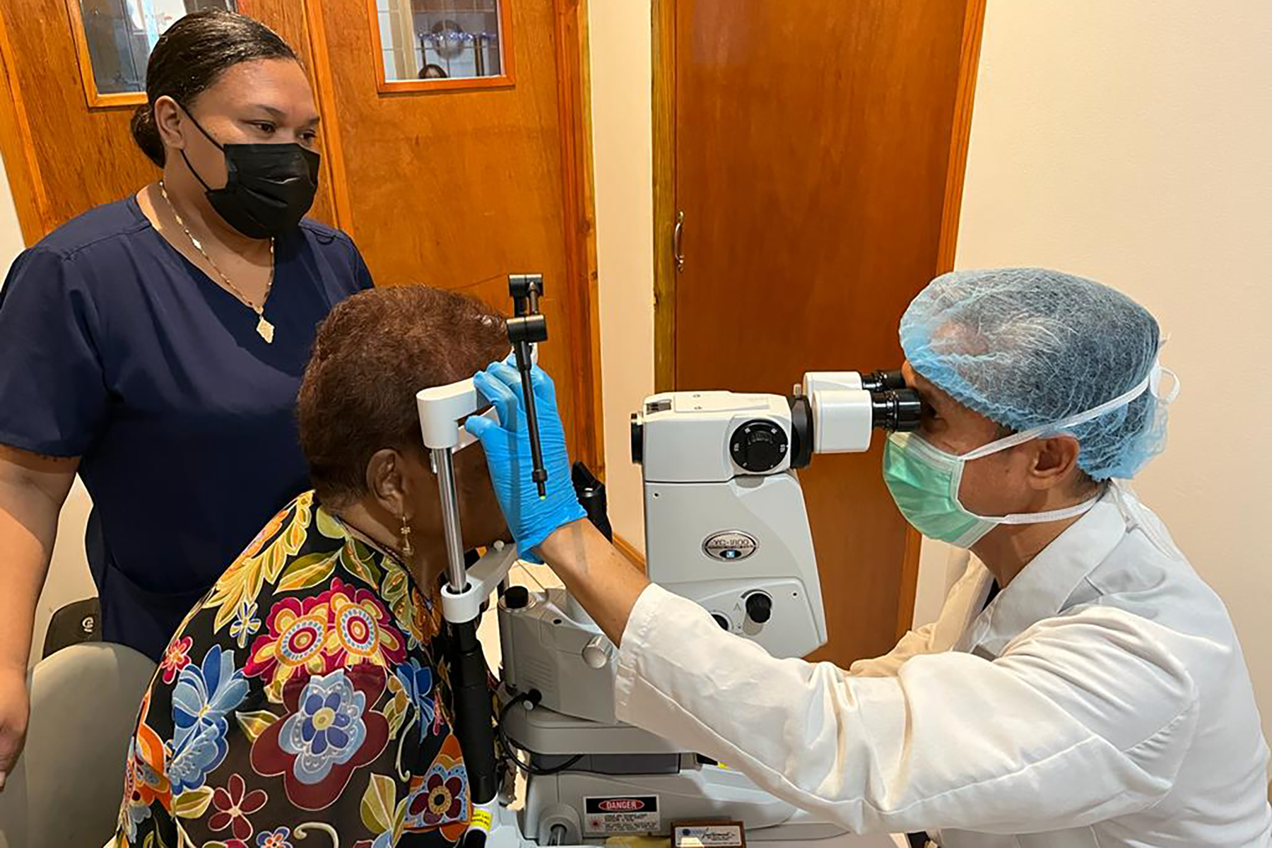 A woman in nursing uniform stands by while a man in doctor's coat gives another woman an eye exam