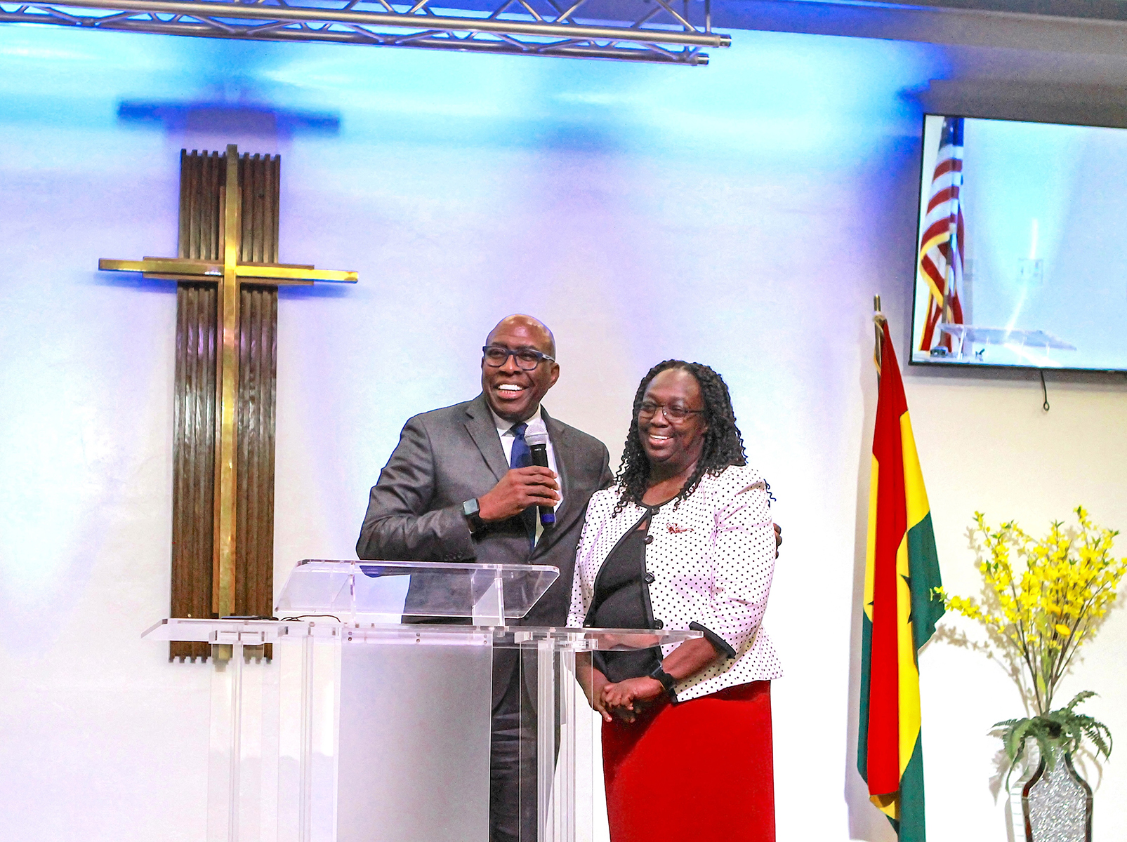 Black couple speak on a podium with cross to the left and Ghanaian flag to the right