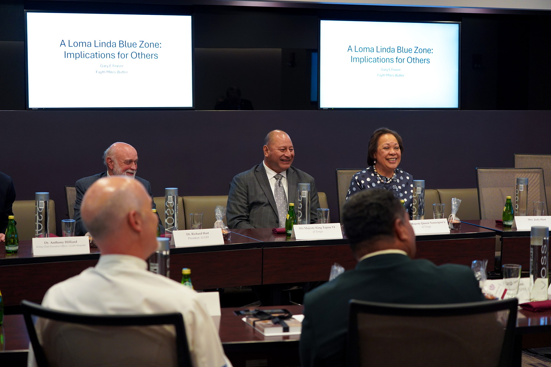 A phofo of a smiling king and queen of Tonga sitting at a boardroom table in front of a slide on the Loma Linda Blue Zone.  